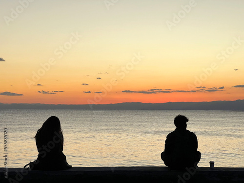 Fotografiet 夕暮れの海を眺めて離れて座る男女のシルエット