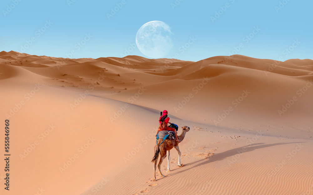A woman in a red turban riding a camel across the thin sand dunes of the in Western Sahara Desert with full moon - Morocco, Africa 