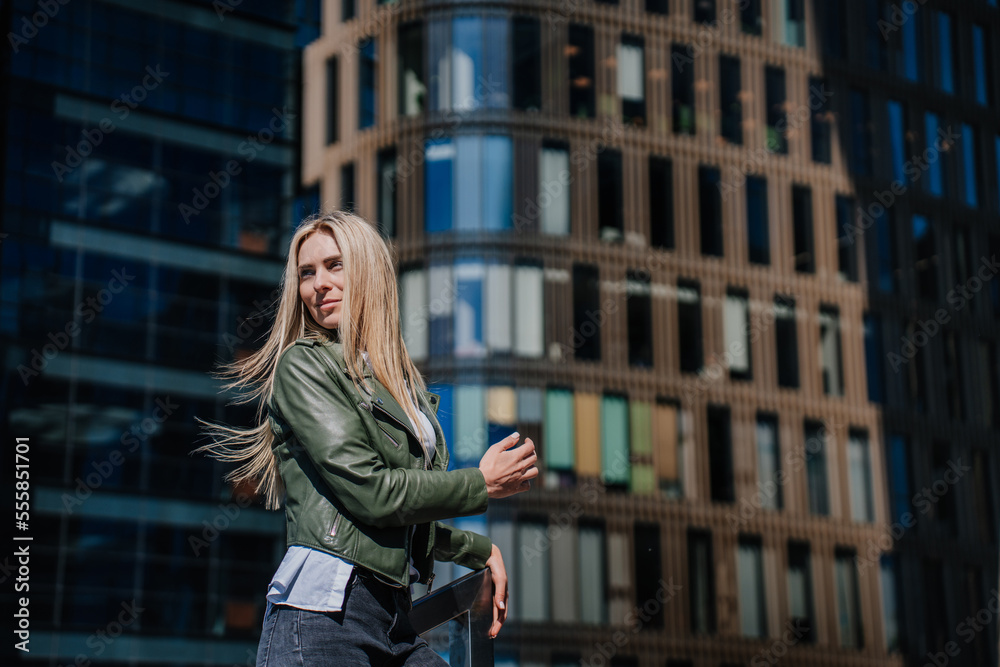 Fototapeta premium Beautiful hipster female standing on chairs outside against business center enjoys sunny day. Attractive young woman tired, walking, waiting for boyfriend. City and people. Perplexed female.