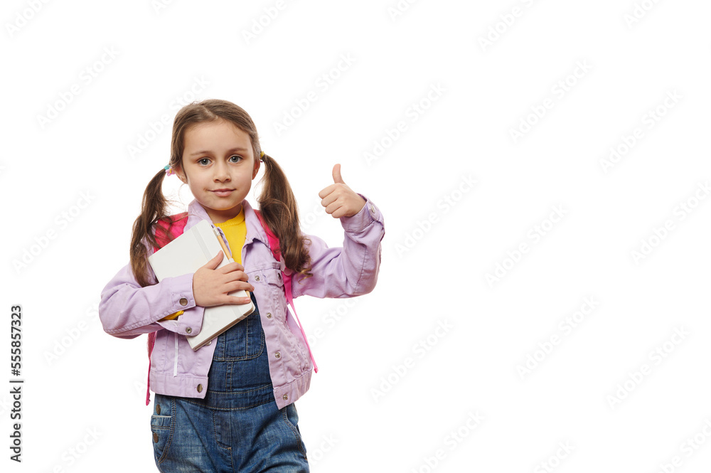Caucasian baby girl, smart first grade schoolgirl in casual denim, holding textbook and showing thumb up, looking at camera, isolated on white background. Back to school on new semester academic year Caucasian baby girl, smart first grade schoolgirl in casual denim, holding textbook and showing thumb up, looking at camera, isolated on white background. Back to school on new semester academic year