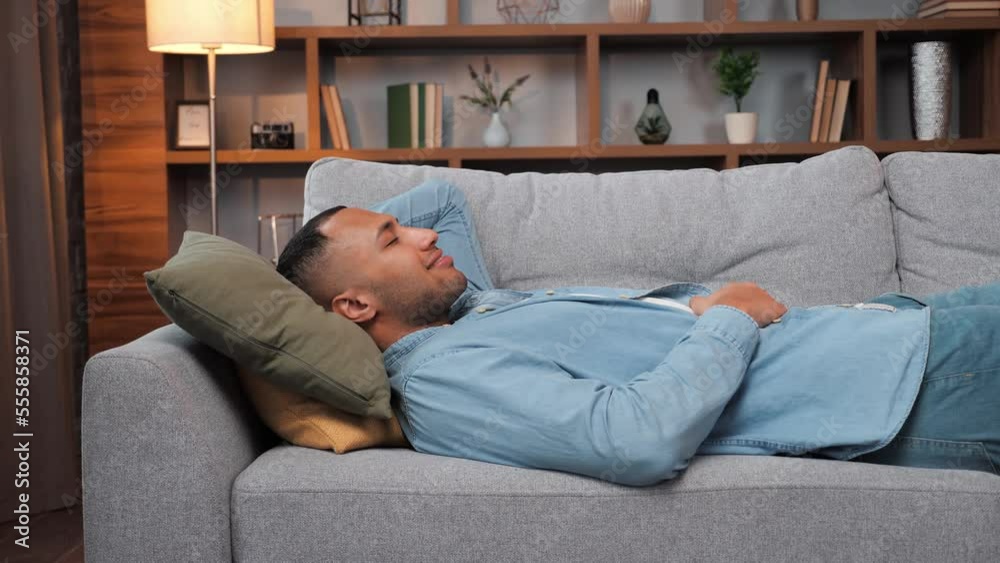 Happy young man laying on sofa, relaxed african american guy resting napping on sofa with eyes closed at home, healthy guy breathing fresh air lounge on couch hands behind head enjoy stress free.