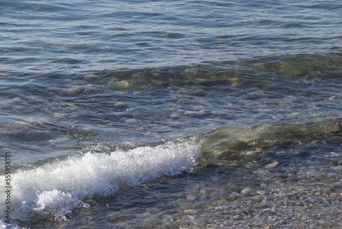 the waves crashing on the shore lined with shells and round stones, the sun's rays shining on the waves, and the ships in the distance