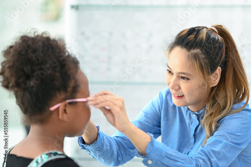 Smiling female optician helping child girl choosing the most appropriate eyeglasses. Eyesight and vision concept