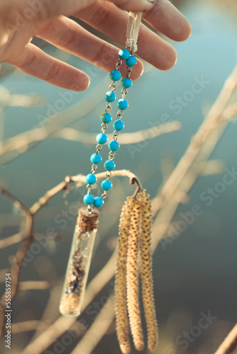 Close up female hand holding necklace with small bottle concept photo. First view person photography with birch twigs on background. High quality picture for wallpaper, travel blog, magazine, article
