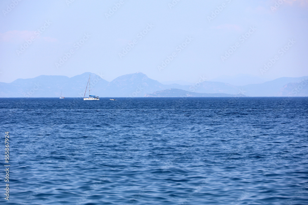 Picturesque view to blue sea with sailing yachts and mountain islands on horizon in mist. Calm water surface, background for traveling and vacation
