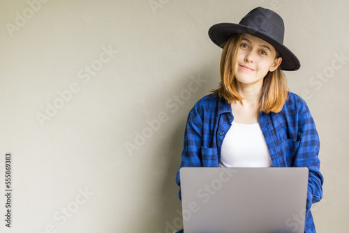 Happy hipster young female wear a hat sitting in the floor working on laptop. Portrait of creative young smiling woman using computer notebook. Beautiful girl sitting on the floor near gray wall.
