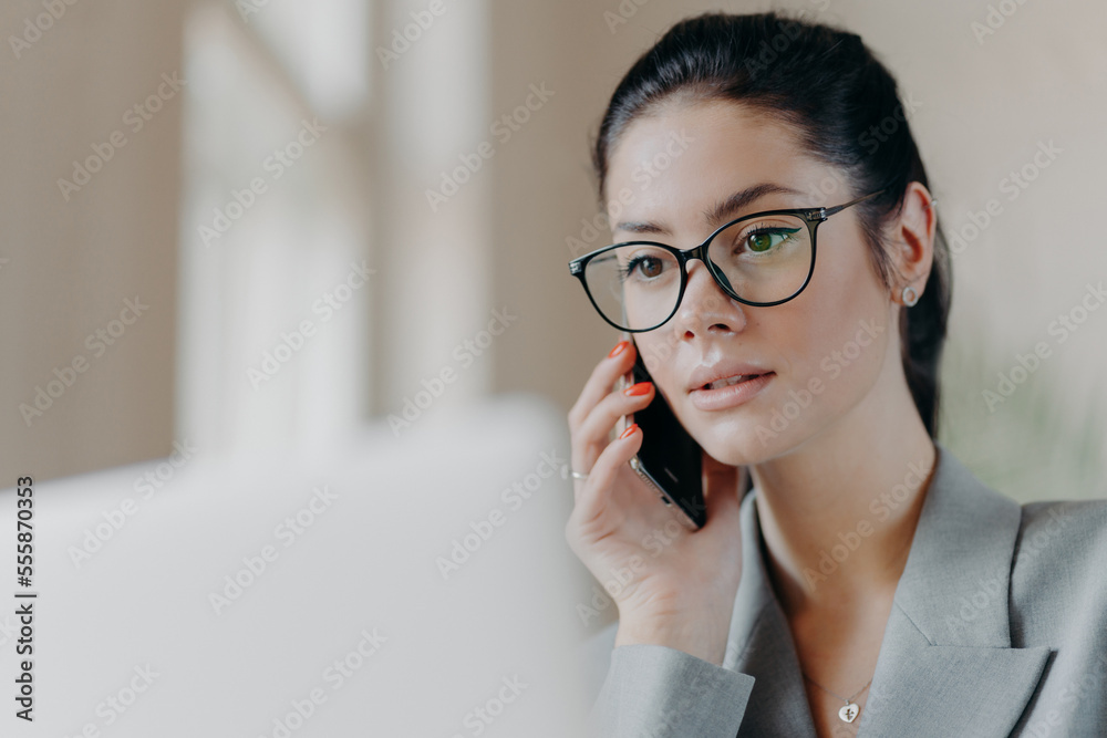 Close up shot of pleasant looking brunette European woman wears transparent glasses, focused in laptop screen, has telephone conversation during remote work, works on project. Technology concept Close up shot of pleasant looking brunette European woman wears transparent glasses, focused in laptop screen, has telephone conversation during remote work, works on project. Technology concept