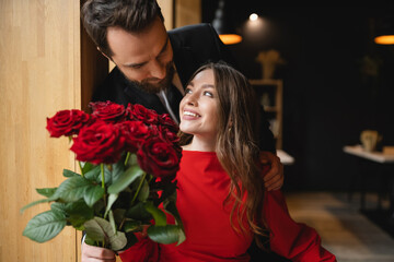 bearded man in suit holding bouquet of red roses near joyful girlfriend on valentines day