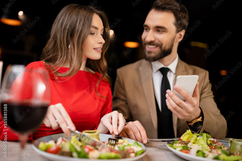 bearded man using smartphone near happy girlfriend in restaurant during celebration on valentines day bearded man using smartphone near happy girlfriend in restaurant during celebration on valentines day