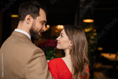 side view of happy and bearded man looking at pretty girlfriend in red dress on valentines day
