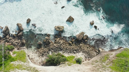 Aerial top down view of a rocky coastline with white foamy waves crashing into the shore. Tall seaside cliff covered with green bush and trees. Camera moving towards the beach, rotate to right. 4k. photo