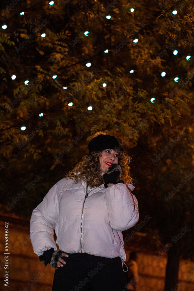 Mujer de raza caucásica con el pelo rizado y gorro de lana disfrutando como una niña pequeña bajo las luces de Navidad en el centro de Madrid.