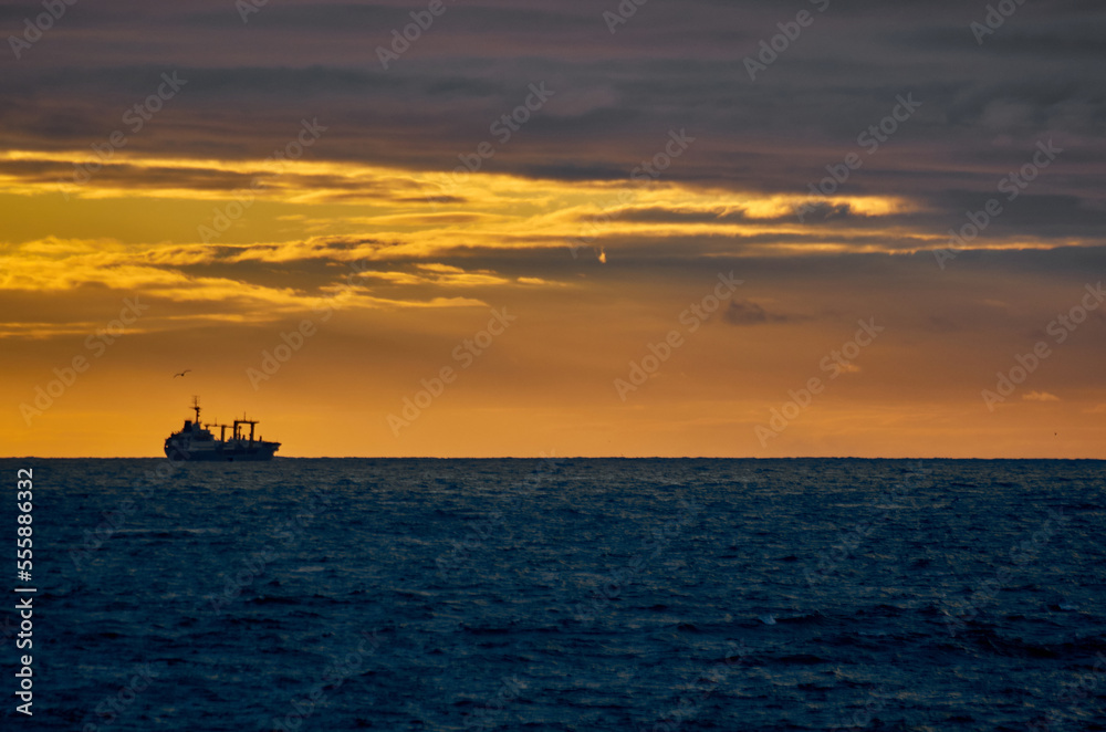 Silhouette of a fishing ship on the horizon of the sea against the backdrop of a dramatic sunset with sunbeams