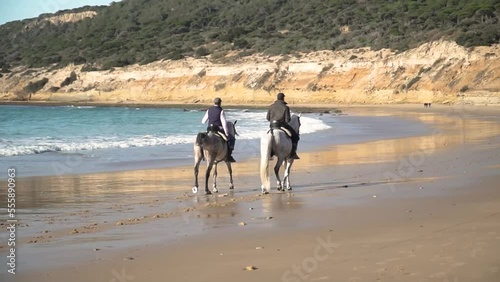 Wide handheld shot of two riders with their horses galloping along the beach with calm waves overlooking the landscape