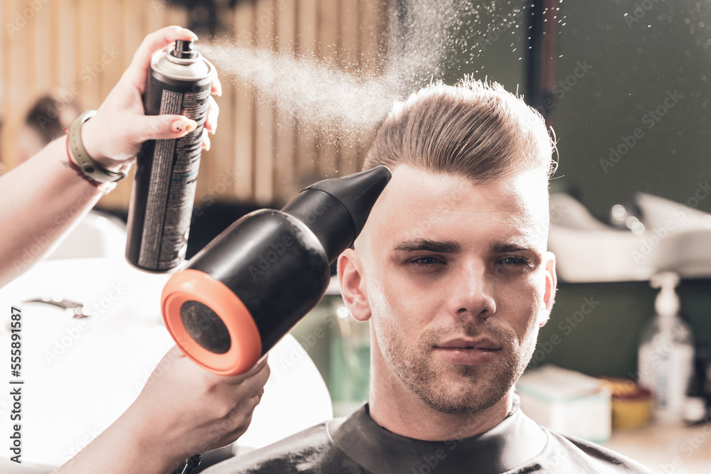 Naklejka premium Girl hairdresser sprays spray and dries hair with a hairdryer to a young guy sitting in a chair in a barbershop