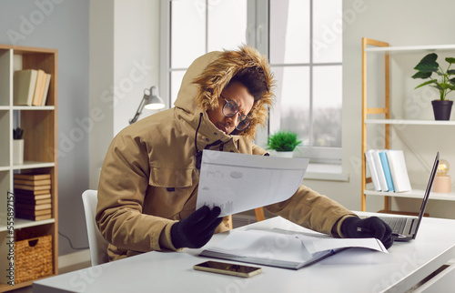African American man in a warm winter jacket and gloves sitting at his desk with a laptop in a cold room at home, looking at heating bills, and comparing costs in different time periods