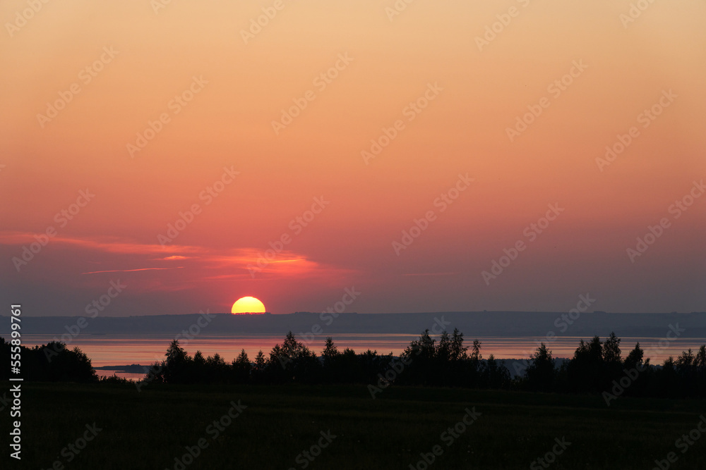 A picturesque summer sunset over the river. The sun hides behind the horizon, painting the sky and water in golden and pink shades. The river is framed by black silhouettes of trees. A picturesque summer sunset over the river. The sun hides behind the horizon, painting the sky and water in golden and pink shades. The river is framed by black silhouettes of trees.