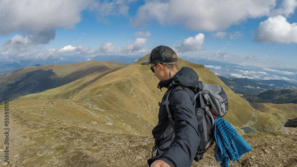 Man with backpack and hat taking selfies with panoramic view of the mountain ranges of Zirbitzkogel Grebenzen, Seetal Alps, Styria (Steiermark), Austria, Europe. Idyllic hiking trails in sunny summer Man with backpack and hat taking selfies with panoramic view of the mountain ranges of Zirbitzkogel Grebenzen, Seetal Alps, Styria (Steiermark), Austria, Europe. Idyllic hiking trails in sunny summer