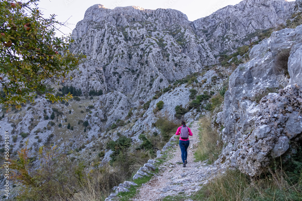Woman with backpack on idyllic hiking trail from Kotor to Derinski Vrh, Lovcen mountains, Dinaric Alps, Montenegro, Balkan Peninsula, Europe. Path is surrounded by steep cliffs and rock formations