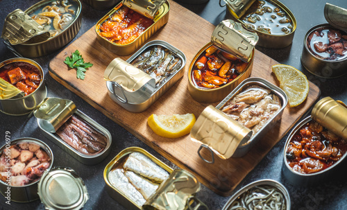 Fotografie Preserve cans with different products, fish and seafood, natural or pickled, open on a dark table with a wooden cutting board and lemon