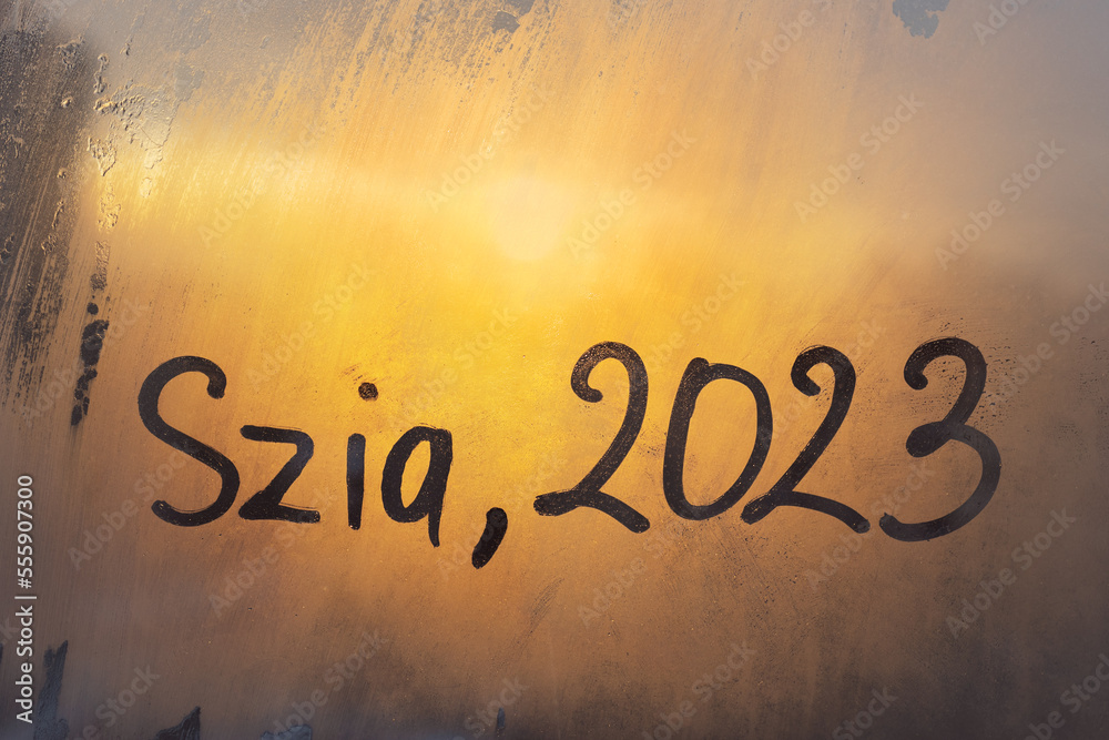 Fototapeta premium lettering Szia in Hungarian is hello in english and numbers 2023 of new year paint with finger on splashed by rain foggy glass on orange sunset frost window