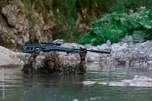 A military man or airsoft player in a camouflage suit sneaking the river and aims from a sniper rifle to the side or to target. © .shock