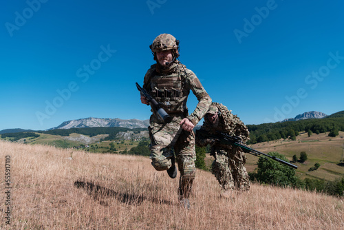 A sniper team squad of soldiers is going undercover. Sniper assistant and team leader walking and aiming in nature with yellow grass and blue sky. Tactical camouflage uniform.