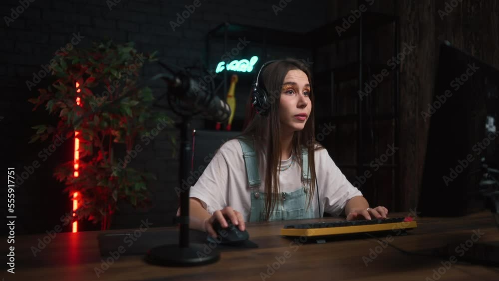 Calm beautiful gamer girl in a headset plays online games on a computer and looks at the camera with a serious face. Calm beautiful gamer girl in a headset plays online games on a computer and looks at the camera with a serious face.