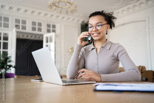 A confident woman with glasses is talking on the phone, smiling, a freelancer is writing a message at the workplace in the office, a laptop is on the table for online work on a startup.