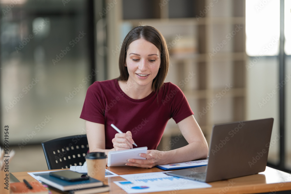 Pretty young businesswoman working on a laptop and taking notes In the office. Pretty young businesswoman working on a laptop and taking notes In the office.