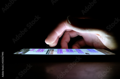 Hand using an app on a smartphone in the dark late at night with lighting from phone screen. Person discreetly text messaging or reading private or personal messages on a cellphone in the darkness.