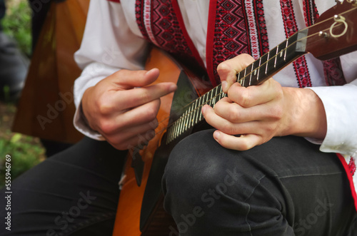 School boy in a white embroidered shirt and black trousers plays a musical instrument balalaika at a concert in the park