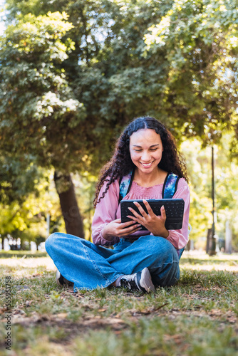 Black university student woman smiling and using a tablet sitting outside in a park on the grass