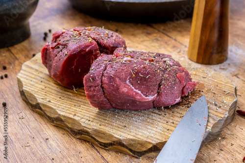 Preparation of raw beef fillet steak on wooden board with steel pan, mortar salt and pepper shakerand rosemary and chilly photographed from the side.