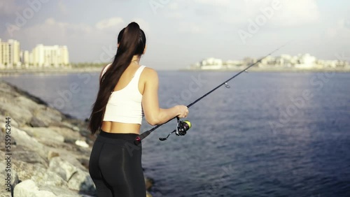 The beginner fisherman girl is casting and fishing on the rocks in the sea. Throws the bait with spinning. Rotate the reel. Rocks and low rise buildings on the background photo