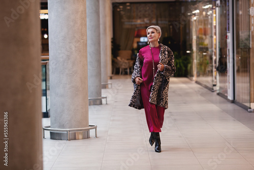 Beautiful middle aged Woman Wearing Fashionable Clothes long burgundy silk dress, leopard print coat jacket, black boots. Female stylish Model walking mall shopping center. Fashion outfit