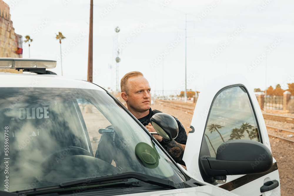 Horizontal image of white male caucasian police officer standing in the door opening of his trooper car with lights on looking away from camera next to railroad tracks.