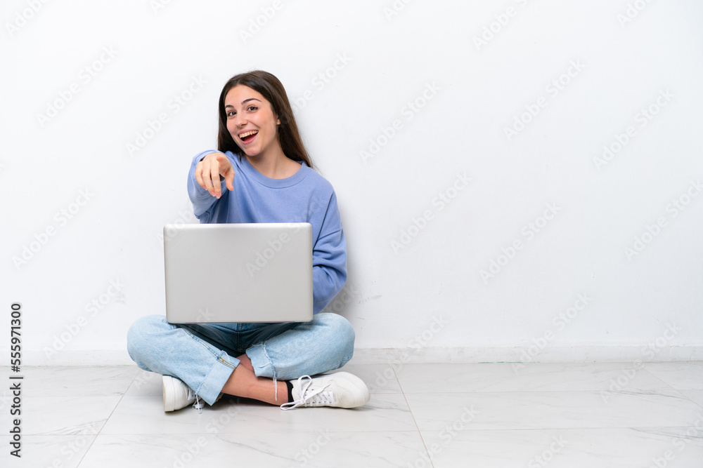 Young caucasian woman with laptop sitting on the floor isolated on white background points finger at you with a confident expression Young caucasian woman with laptop sitting on the floor isolated on white background points finger at you with a confident expression