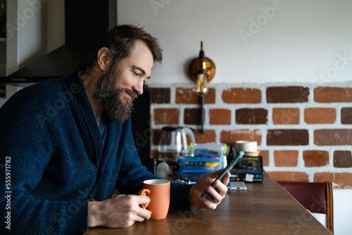 Shot of a handsome young man drinking coffee at home. Happy man drinking a cup of coffee at home. Shot of a young man using a smartphone and having coffee in kitchen at home.