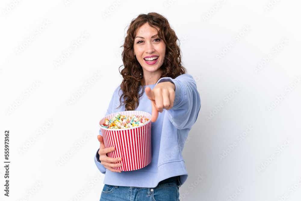 Young woman with curly hair isolated on white background holding a big bucket of popcorns while pointing front