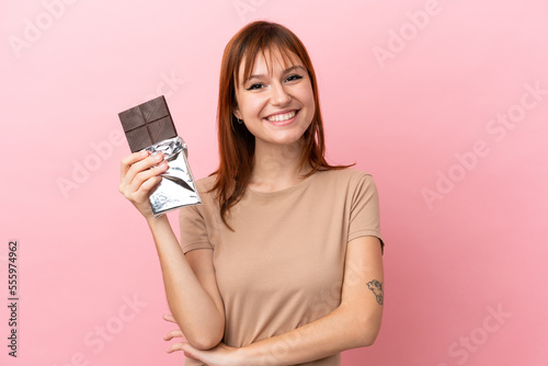 Redhead girl isolated on pink background taking a chocolate tablet and happy