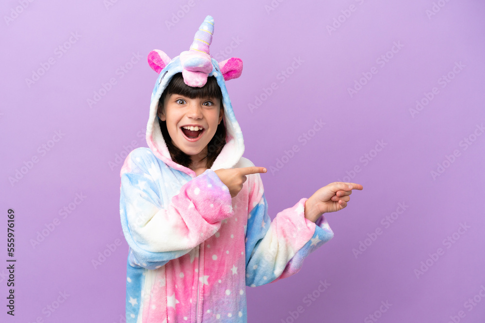 Little kid wearing a unicorn pajama isolated on purple background surprised and pointing side