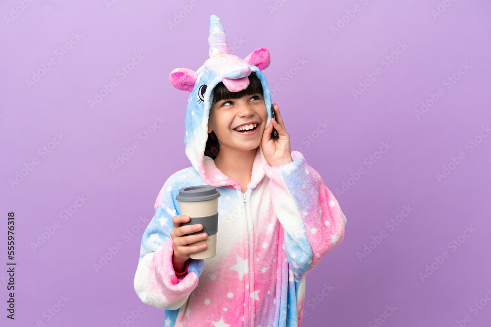 Little kid wearing a unicorn pajama isolated on purple background holding coffee to take away and a mobile Little kid wearing a unicorn pajama isolated on purple background holding coffee to take away and a mobile