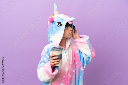 Little kid wearing a unicorn pajama isolated on purple background holding coffee to take away and a mobile