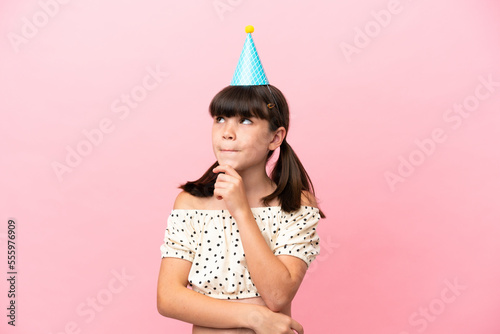 Little caucasian kid with birthday hat isolated on pink background having doubts and thinking