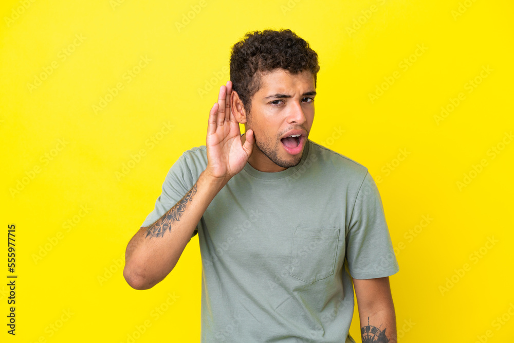 Young handsome Brazilian man isolated on yellow background listening to something by putting hand on the ear Young handsome Brazilian man isolated on yellow background listening to something by putting hand on the ear