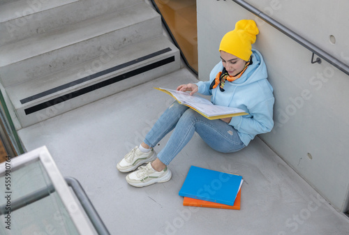 A female student in a blue sweatshirt and yellow cap sits on a stone floor between stairs waiting for class photo