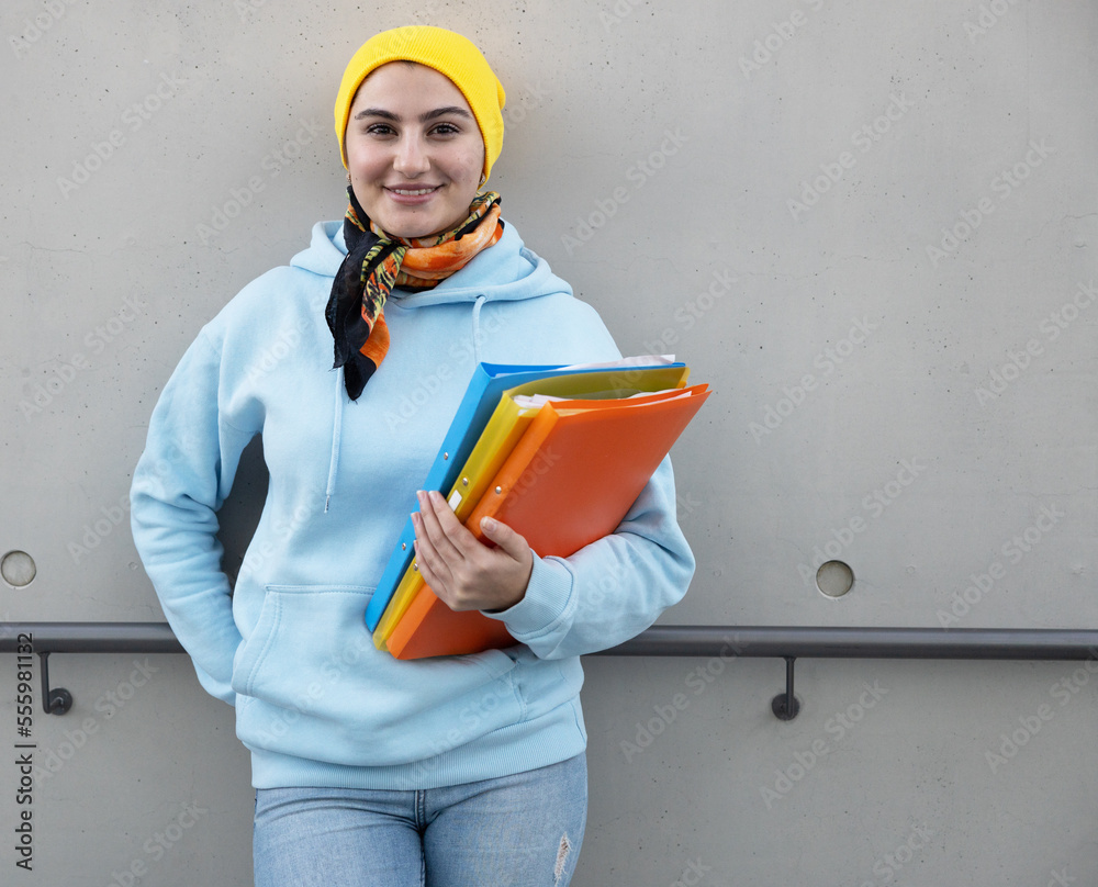 Smiling female student in a blue sweatshirt and yellow cap stands with folders near the wall