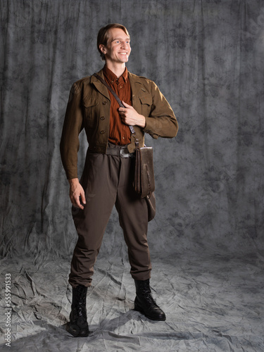 A young guy in military-style clothes, a brown flight jacket and breeches with suspenders. Posing in the studio on a gray background