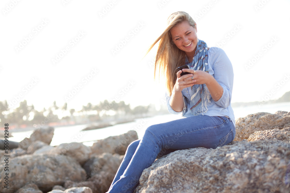 Young Woman Sitting on Rocks and Texting on Cell Phone at Beach, Jupiter, Palm Beach County, Florida, USA Young Woman Sitting on Rocks and Texting on Cell Phone at Beach, Jupiter, Palm Beach County, Florida, USA